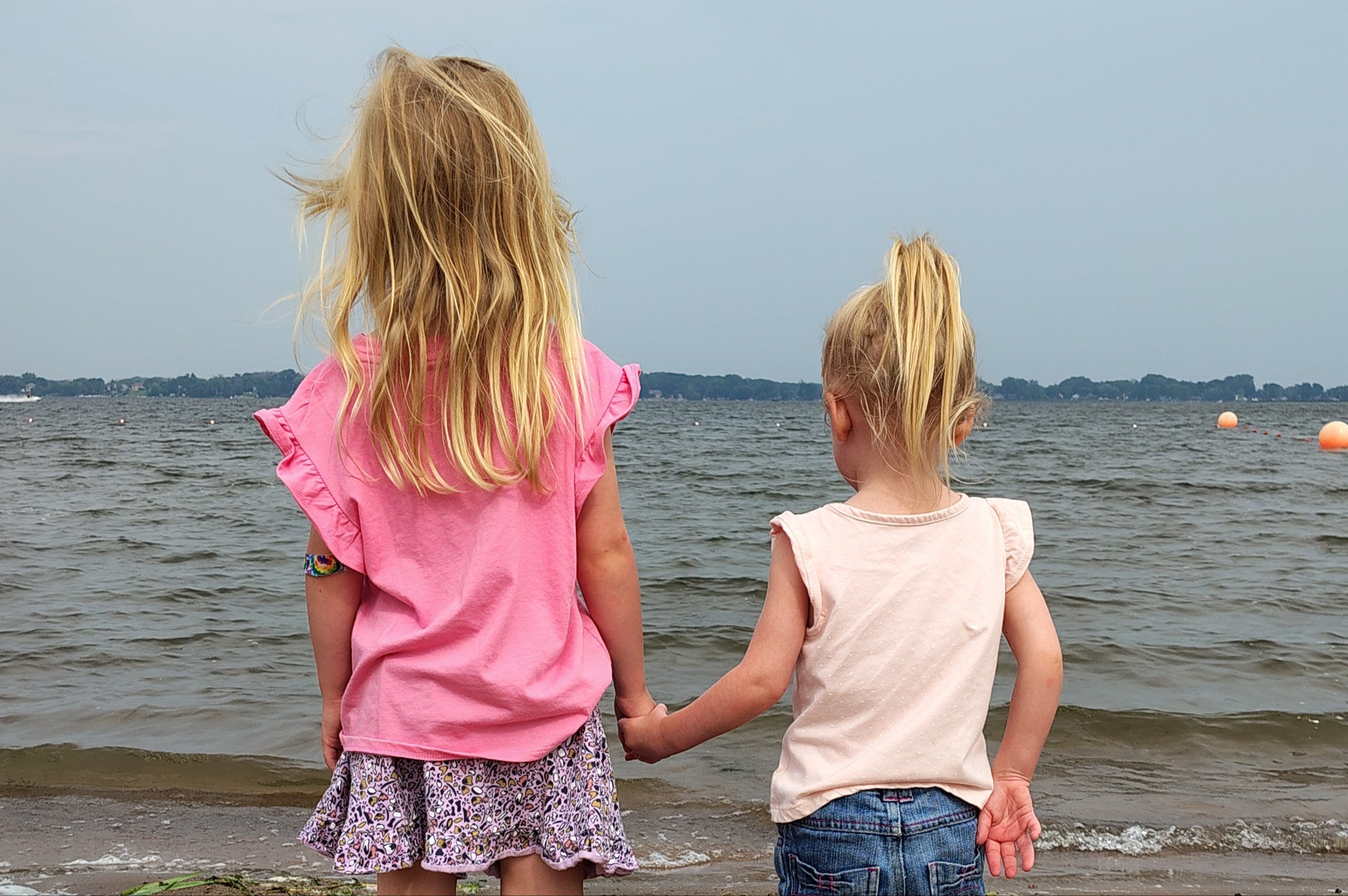 Two young girls holding hands on a beach with water and sky in the background.